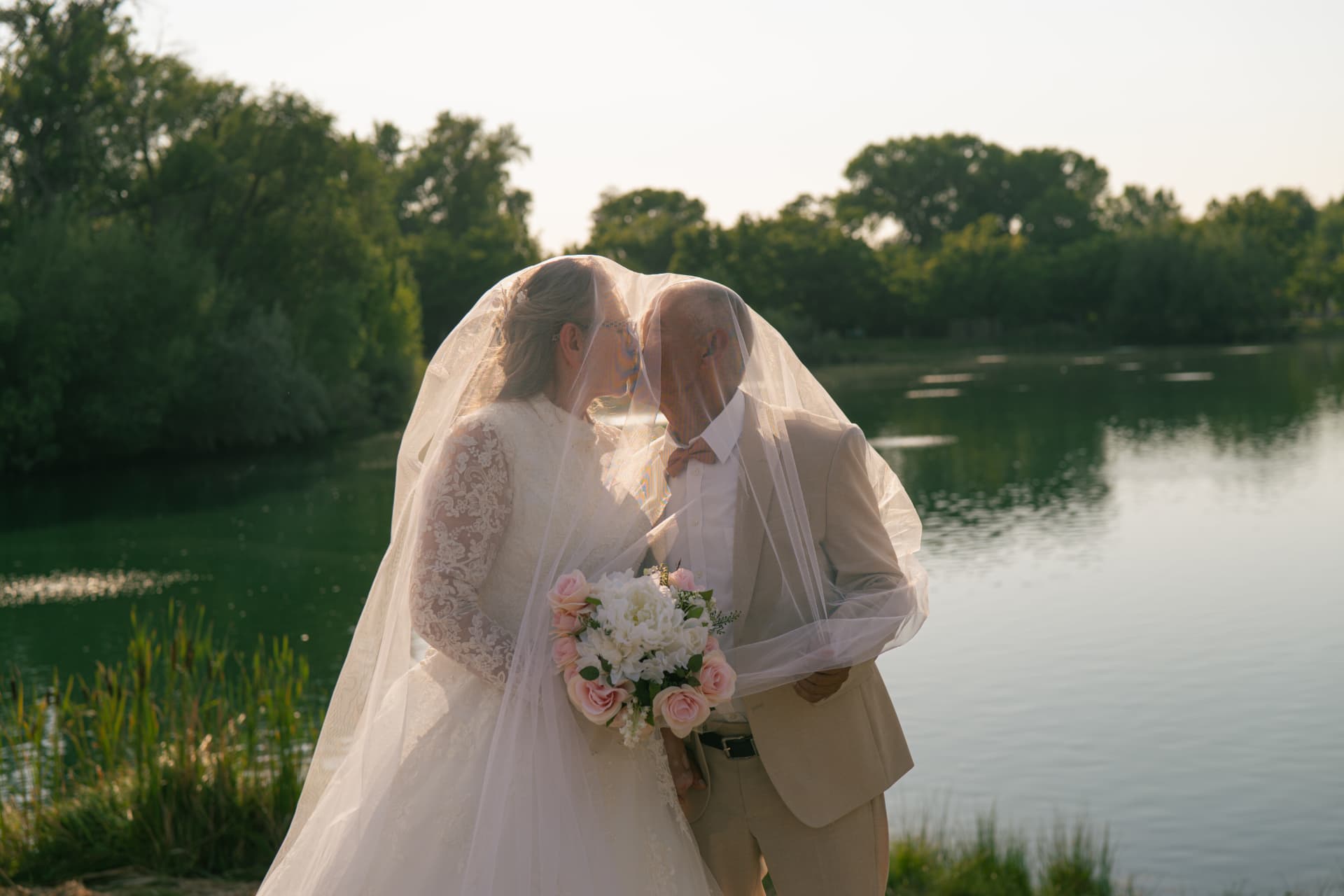 Bride and groom kissing under a veil by a lake